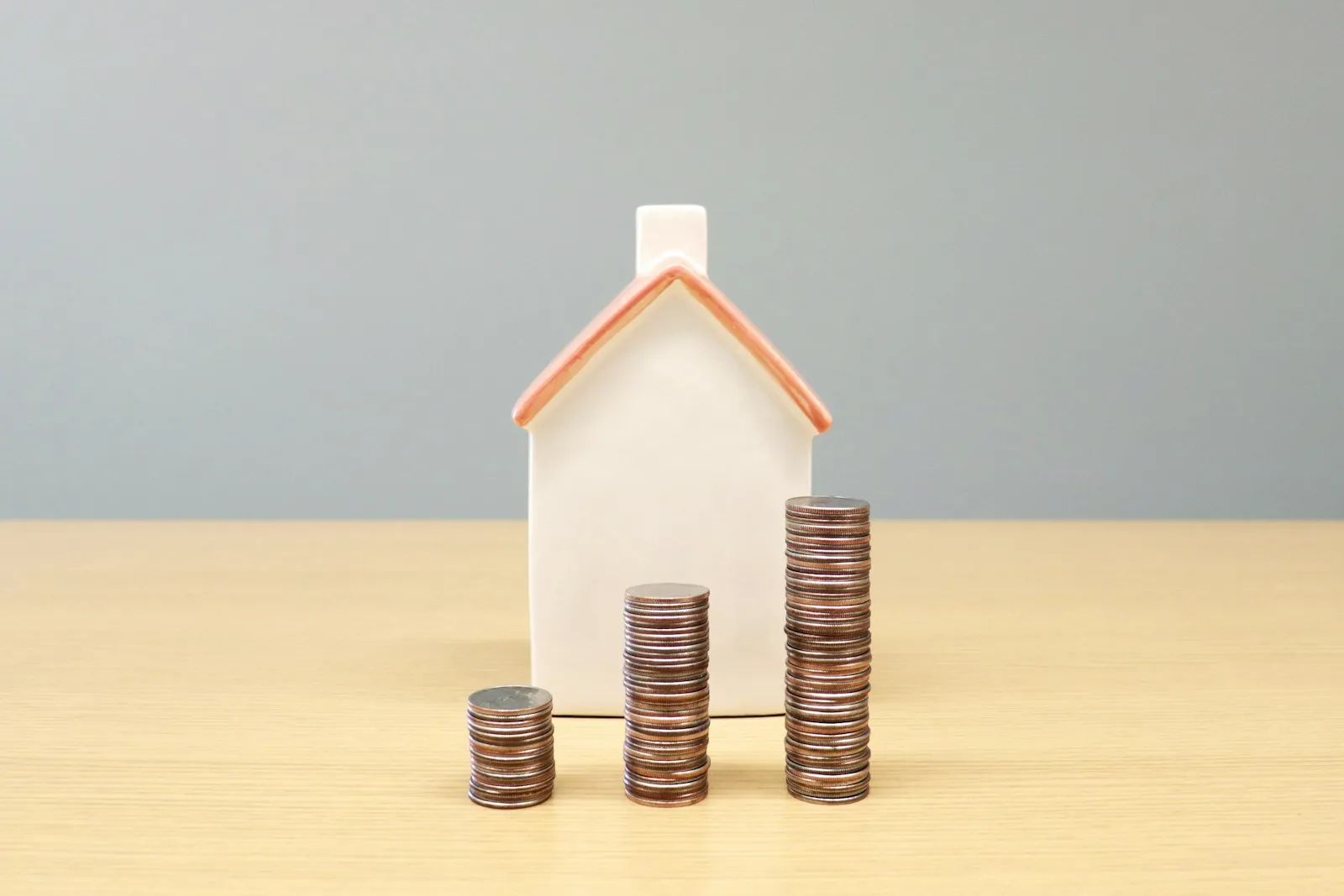 Une maison miniature posée devant des piles de pièces, sur une table claire