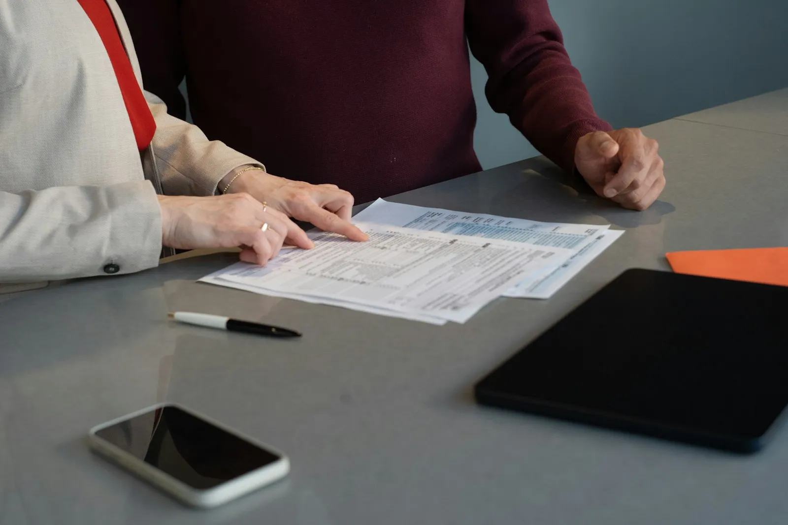 Des mains sur des documents posés sur une table, avec un stylo, un téléphone et une tablette