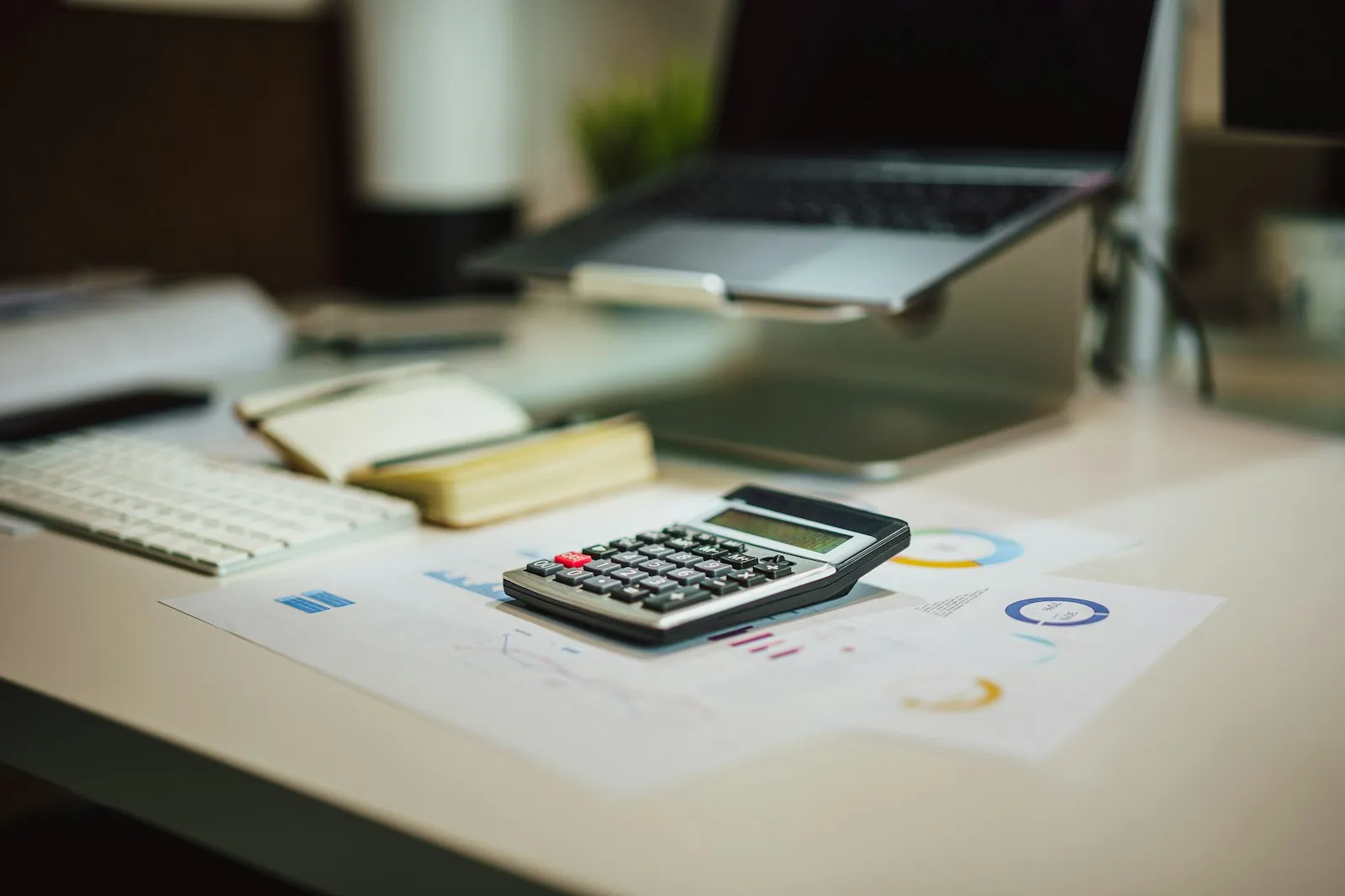 Une calculatrice posée sur des graphiques et un carnet, devant un ordinateur portable, sur un bureau clair