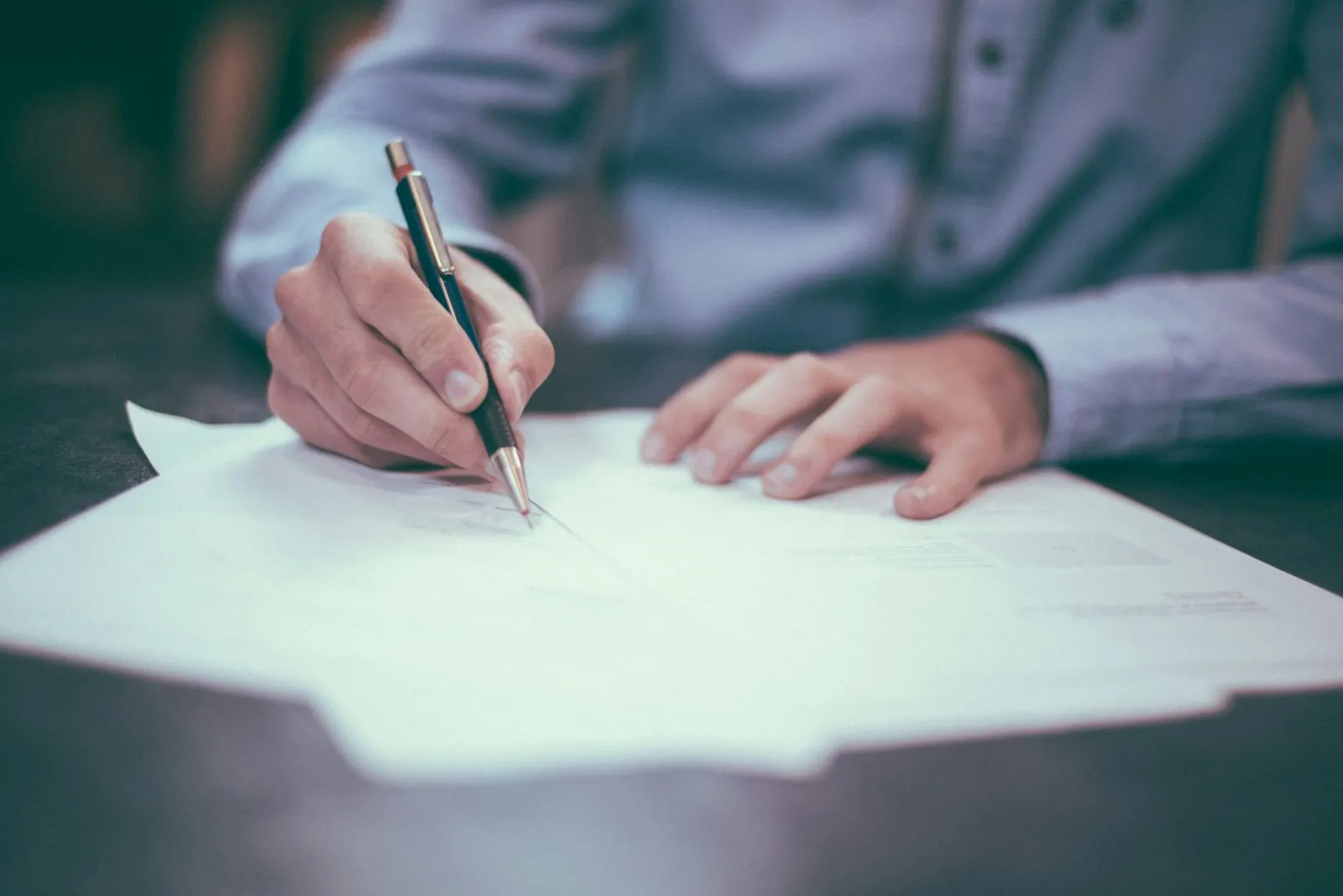 Une main signe un document au stylo, sur une pile de feuilles posée sur une table