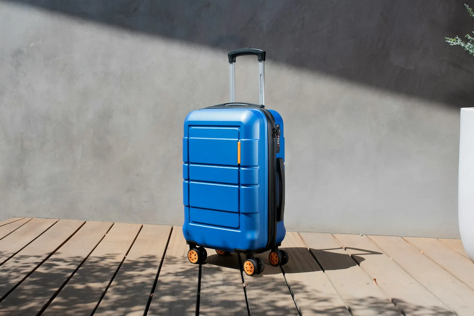 Une valise bleue à roulettes posée sur une terrasse en bois, devant un mur gris