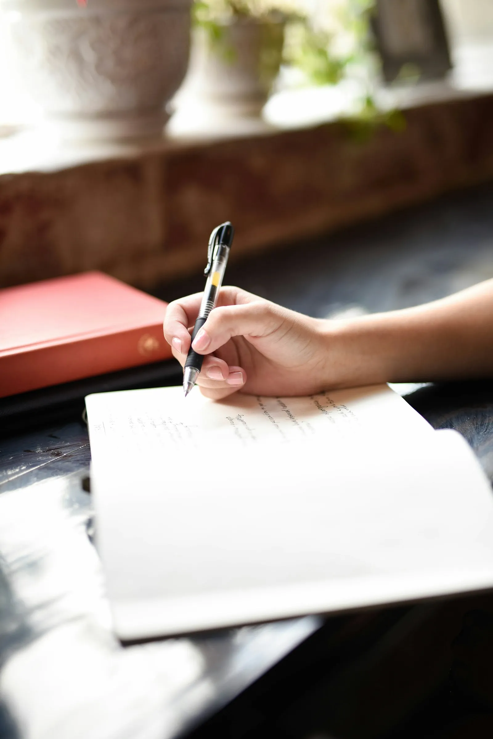 Une main écrit avec un stylo dans un cahier, à côté d'un classeur rouge, près d'une fenêtre