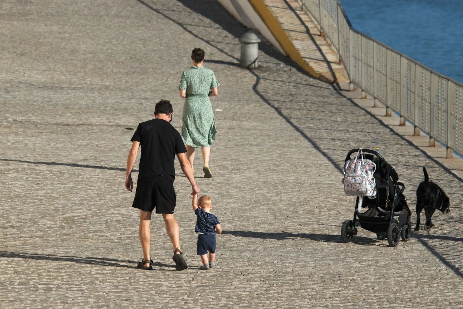 Parents vus de dos sur un quai ensoleillé, le père tenant la main de leur jeune enfant, avec la poussette garée à côté
