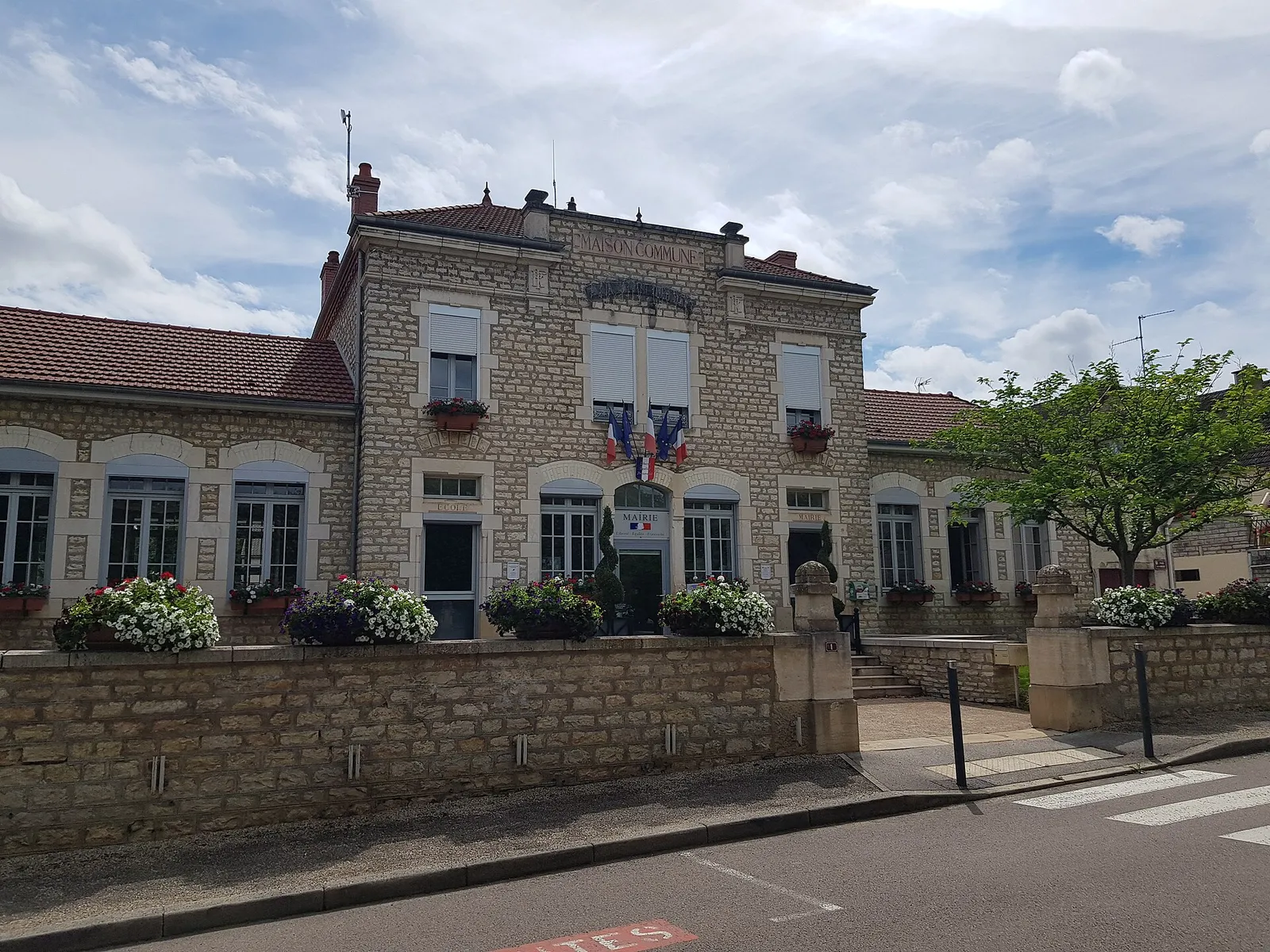 La mairie-école de Dracy-le-Fort, bâtiment en pierre marqué « Maison commune », fleuri, sous un ciel bleu