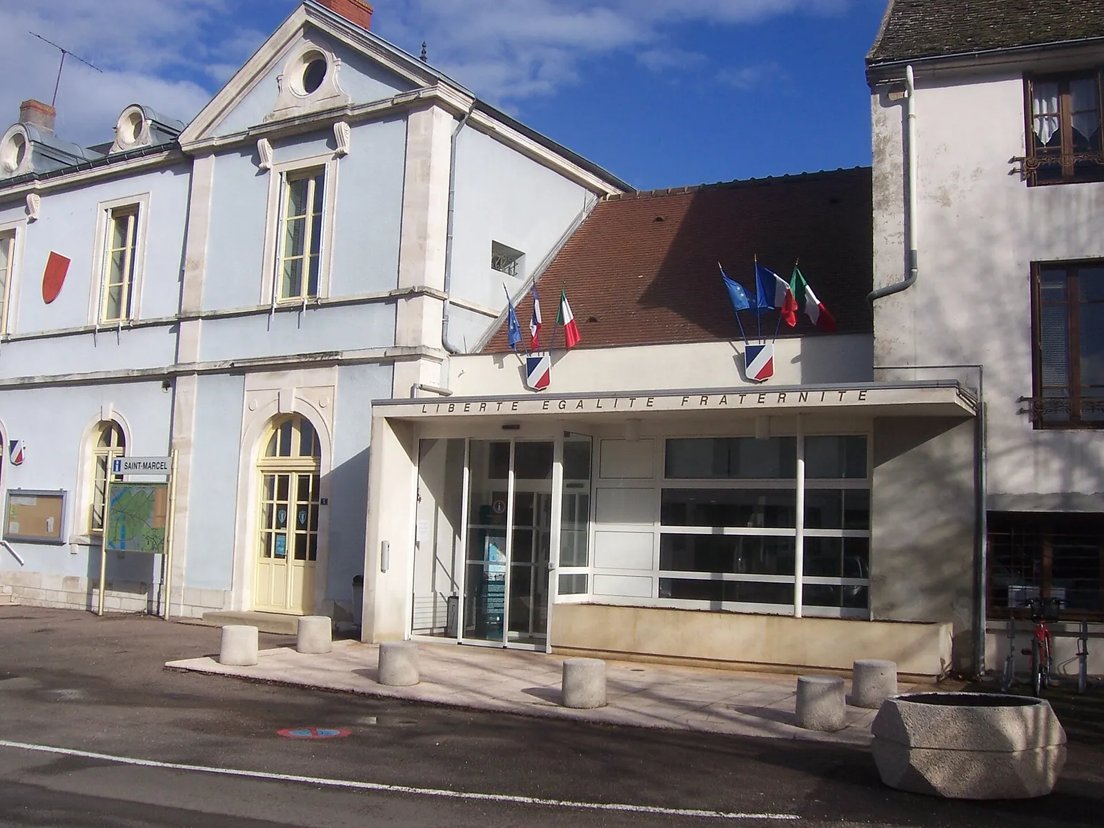 La mairie de Saint-Marcel, façade bleu clair et entrée vitrée pavoisée, sous un ciel bleu